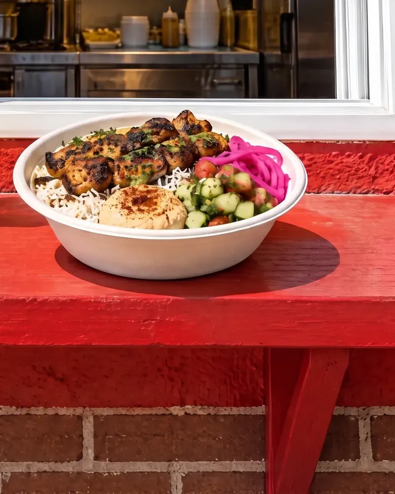 A kebab bowl with rice, hummus, cucumber salad and pink pickled onions, on a red counter.